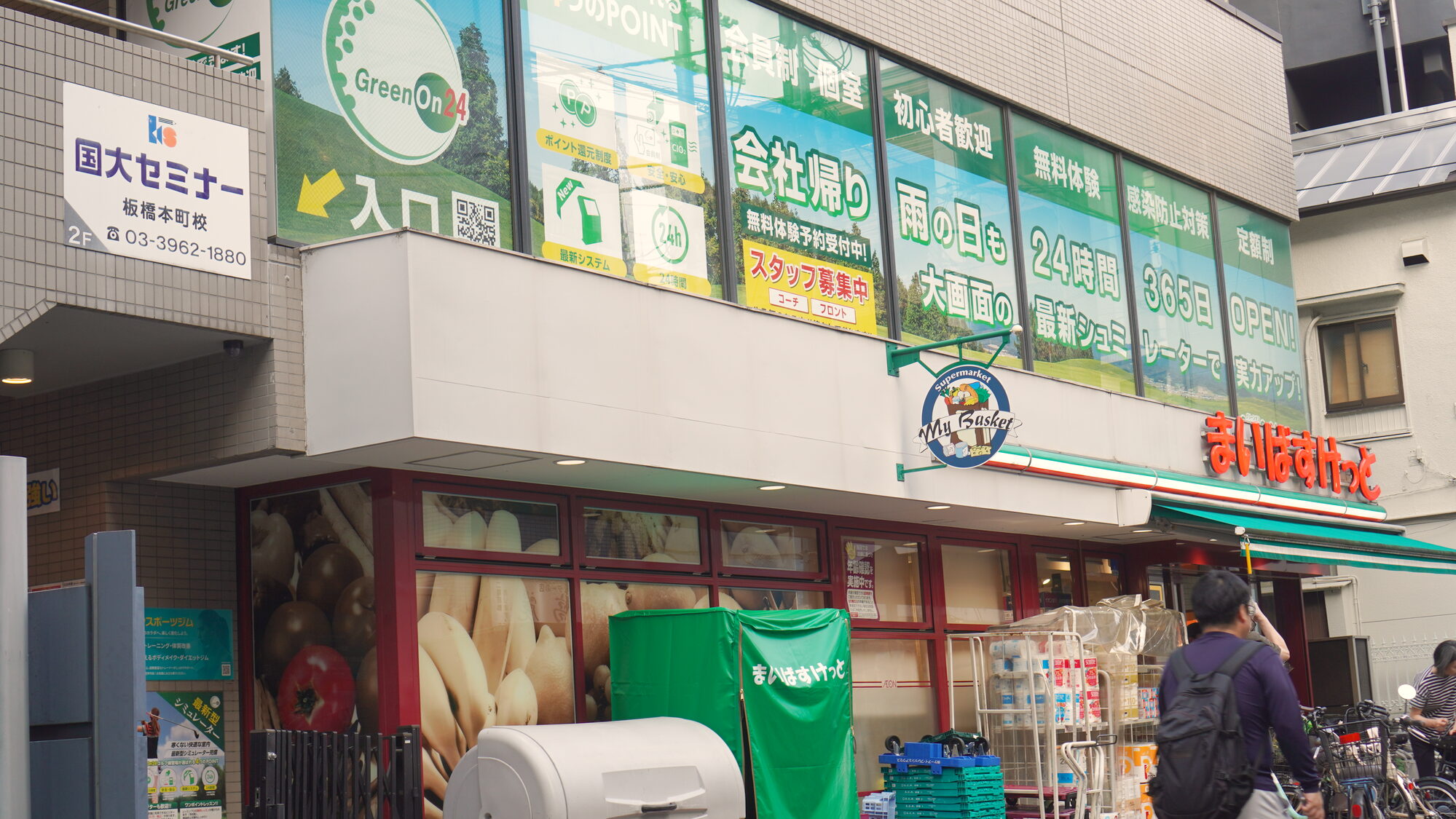 まいばすけっとの建物の外観。2 階の窓に GreenOn24 の看板、1 階に「入口」の案内板があります